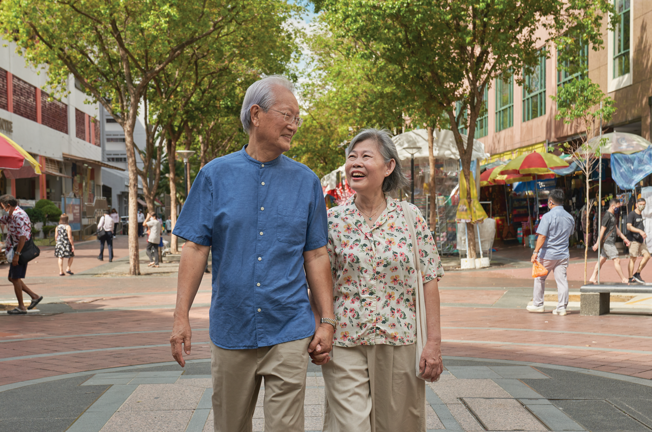 An elderly couple holds hands and smiles as they walk along a tree-lined city street with other pedestrians and market stalls in the background.